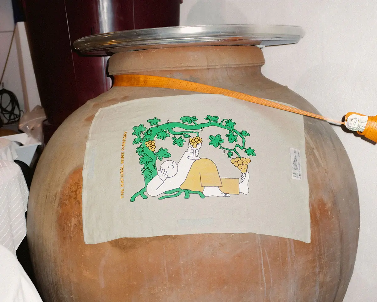 A tea towel designed by Jhon Boy in collaboration with Frama and The Natural Wine Company (TNWC), draped over a large clay wine vessel. The illustration features a character reclining under grapevines, holding a bunch of grapes, with the text ‘The Natural Wine Company’ above. Photograph by Miguel Triano