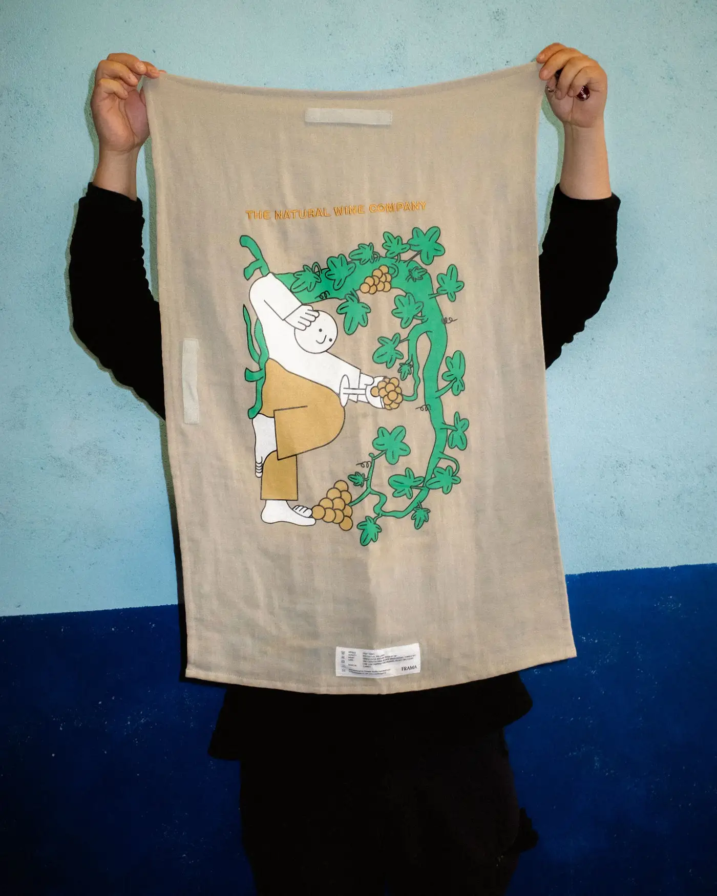 A person holding up a tea towel designed by Jhon Boy in collaboration with Frama and The Natural Wine Company (TNWC). The towel showcases an illustration of a whimsical character harvesting grapes amidst lush grapevines, with the text ‘The Natural Wine Company’ at the top. Photograph by Miguel Triano.