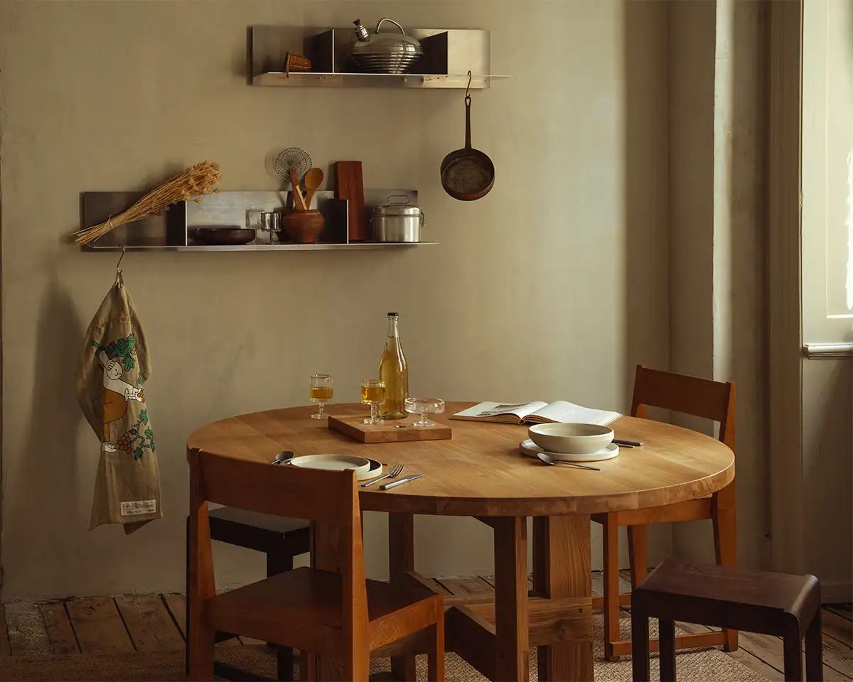 A cozy kitchen setup featuring wooden furniture and shelves adorned with kitchen essentials. A tea towel hangs on the wall with an illustration by Jhon Boy, created in collaboration with Frama and The Natural Wine Company (TNWC). The tea towel features a playful character surrounded by grapevines. Photograph by Miguel Triano.