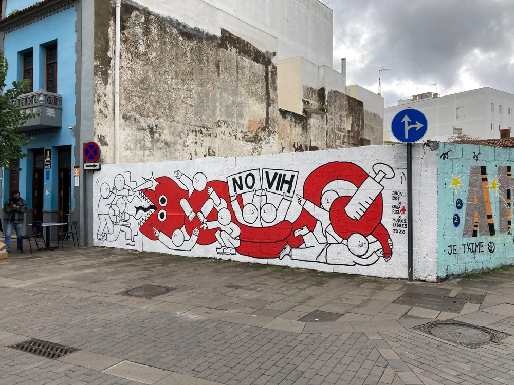 A vibrant mural on a street wall showcasing a cartoonish red creature being tackled by groups of people in white with dynamic expressions, holding signs and objects to subdue it. The central text reads ‘NO VIH,’ symbolizing a message against HIV/AIDS. The mural is signed ‘Jhon Boy Cruz Roja Nuestros Libres 2020’ and is accompanied by urban street elements.