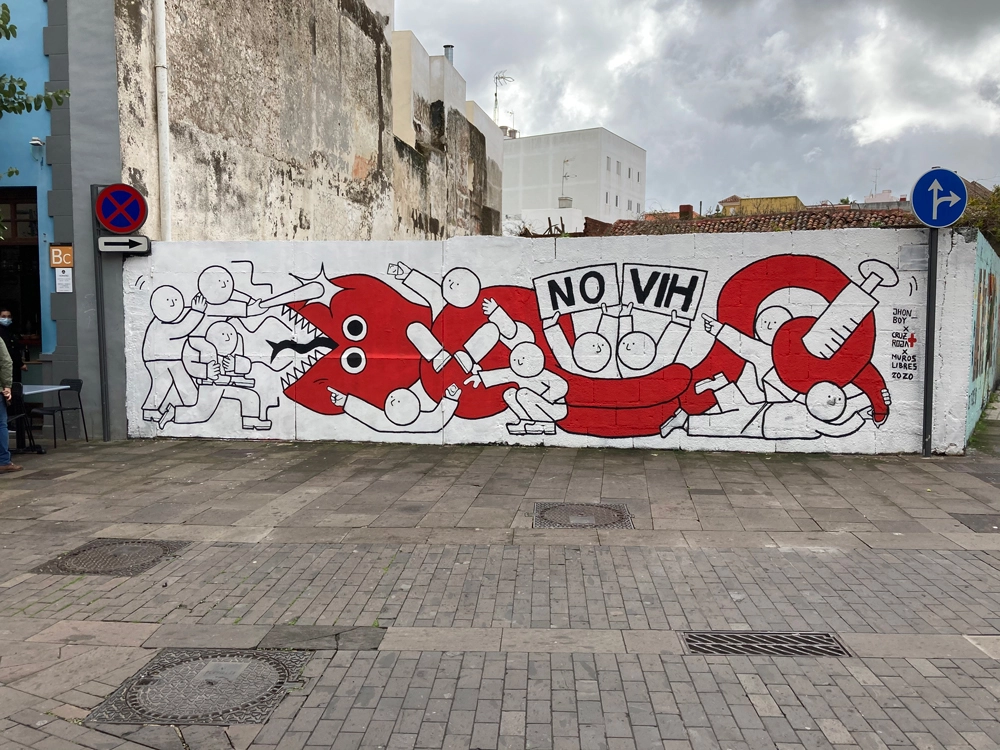 A wide view of a colorful mural located on a street corner, illustrating a red cartoonish creature subdued by people in white with a clear message in bold ‘NO VIH,’ advocating against HIV/AIDS. Additional urban elements such as signs and adjacent artworks can be seen in the surrounding context